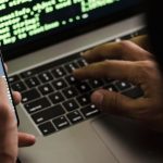Hacks - Unrecognizable hacker with smartphone typing on laptop at desk