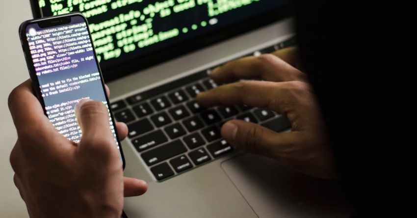 Hacks - Unrecognizable hacker with smartphone typing on laptop at desk