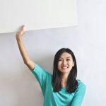 Promotions - Cheerful Asian woman sitting cross legged on floor against white wall in empty apartment and showing white blank banner