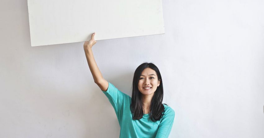 Promotions - Cheerful Asian woman sitting cross legged on floor against white wall in empty apartment and showing white blank banner
