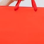 Clearance - Crop unrecognizable female showing red paper shopping bag in hand against white wall