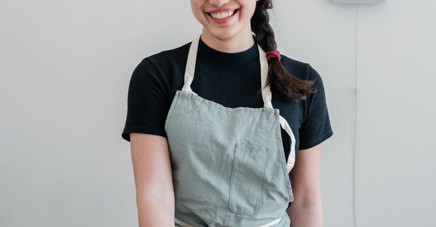 Offers - Positive young Asian woman wearing apron giving paper cup with coffee to go while standing at wooden counter during work in modern coffee house and looking at camera with smile